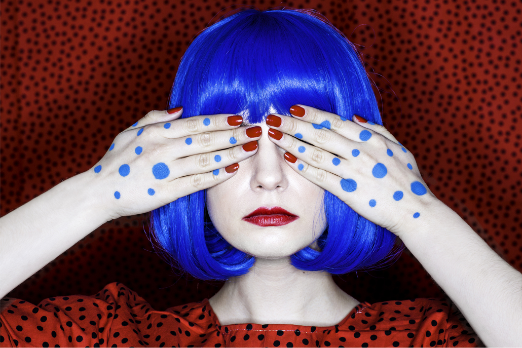 ﻿Woman with blue hair holding hands over eyes with red and black polka-dotted background